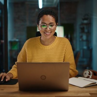 Portrait of Young Latina Marketing Specialist Working on Laptop Computer in Busy Creative Office Environment. Beautiful Diverse Multiethnic Female Project Manager is Browsing Internet. (Portrait of Young Latina Marketing Specialist Working on Laptop C